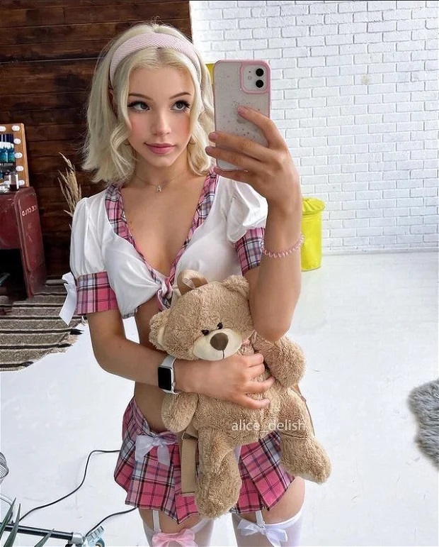 A young blonde with a princess face and small breasts takes a selfie holding a teddy bear